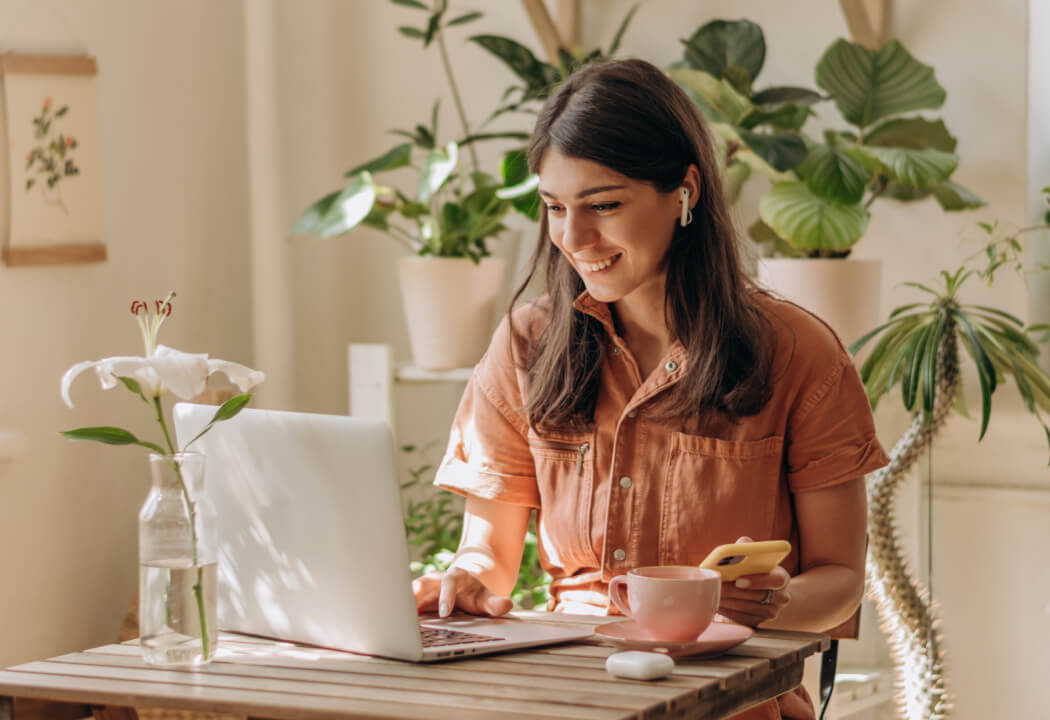 Young woman sitting at a small table with a laptop, smartphone, and coffee cup with house plants surrounding her